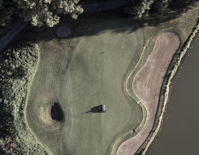 Aerial view of a golf green with a golf cart near the hole, sand bunker on the right, and a water hazard adjacent to the green.